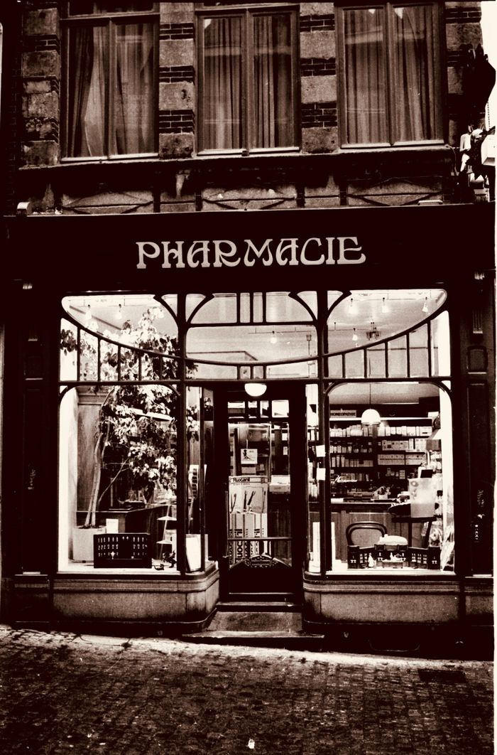 A sepia-toned image of a traditional pharmacy exterior at dusk in Mons, Belgium.